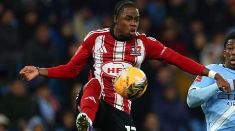 Wigan Athletic player Akeel Higgins, in the red and white of Exeter City last season, tries to control a high ball under challenge from Manchester City player Ryan McAidoo, in sky blue.