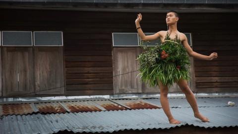 Man dressed in grasses, standing on a tin roof