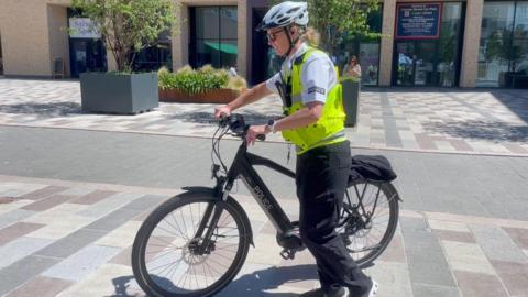 A police officer wearing a high=vis gilet steers a black police bicycle towards the left of the frame. She is wearing a white helmet, black trousers and a white shirt.