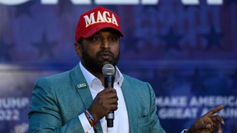 Kash Patel, wearing a red MAGA hat, and speaking into a microphone and pointing