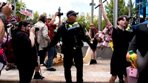 A police officer looks on as protesters wave flags and signs - one saying 'no pride genocide' and a Palestinian flag in view - in Perth on Monday.