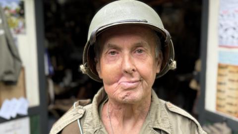 A close up of a man in khaki army fatigues and hard hat. His face is disfigured, with nose and mouth twisted to the right. The open doors of a shed can be seen behind him.
