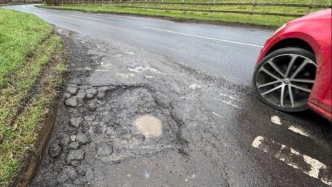 There is a pothole filled with brown rain water at a junction onto a main road. Pieces of the road surface are also broken beside it. A red car is driving past the pothole and the tyre and side of part of the car is in shot.
