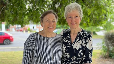 Two women stand together under a green tree. On the left, Angela has short grey hair and is wearing a black and white patterned top, with a silver necklace. On the right, Joan Woodhouse has short white hair, and is wearing black top with white flowers. they have their arms around each other. 