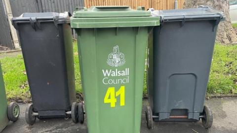 There are two black wheelie bins, on the left and right, with a green one in the middle that includes the words Walsall Council and the number 41. Grass and a fence are behind them.