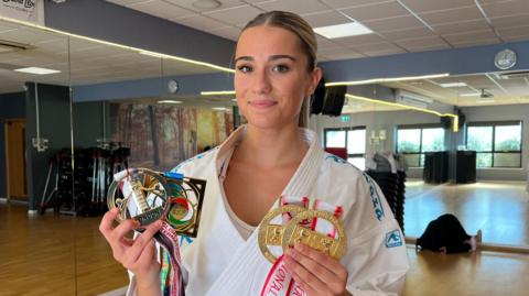 Carla, a young woman with her blonde hair tied back in a ponytail. She is standing in a karate studio which has a wooden laminate floor and a mirror across one wall. She is holding four medals.