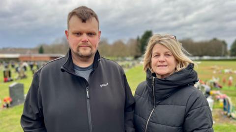 A man with short brown hair and a goatee beard wearing a black zipped jumper is standing next to a woman with short blonde hair and glasses on her head. She is wearing a black coat zipped up and the pair are standing in a crematorium grave yard.