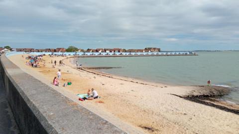 Canvey Island's beach, with the photo taken from behind the concrete sea wall. Adults and children are on the beach. The blue painted sea wall, facing out to sea, can be seen curving round the bay. It is slightly overcast above.