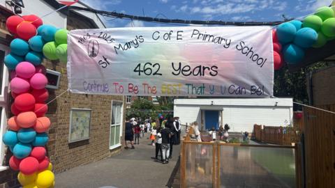 A bright banner at St. Mary's C of E Primary School celebrating 462 years with the message, "Continue To Be The BEST That U Can Be!". Colorful balloons decorate the banner and people are gathered in the background.