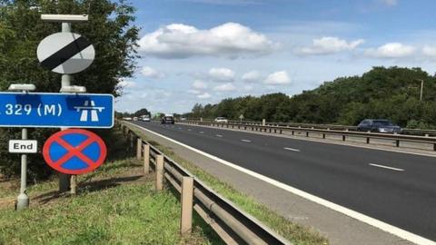 Blue motorway sign to the left with grass verge and barrier at side of dual carriageway and central reservation with vehicles seen beyond