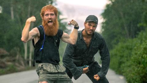 Two men pose, showing off their biceps, in the middle of a road in the jungle.