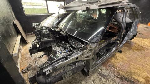 A grey car shell with no doors, wheels or side windows. The car looks like it is ready to be scrapped.