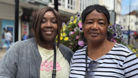 Two women - one in a blue and white striped long sleeved top with a handbag strap over her shoulder and sunglasses hanging off her collar, and the other in a yellow T-shirt and grey jacket with a crossbody bag strap visible, are standing on a high street and smiling at the camera. There are shops, floral displays and people blurred in the background.