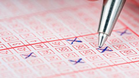 A silver pen hovers over a lottery ticket - some of the numbers already have blue ink Xs through them