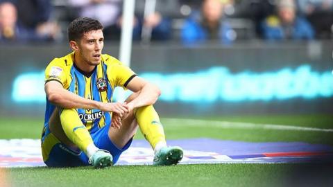 Ross Stewart sittting on the turf with hands on knees wearing Saints yellow and blue striped away kit