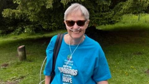 A woman with short grey hair, wearing an oxygen tube, sunglasses and a blue shirt, stood against a green lawn