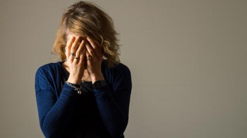 A woman in a blue top stands in front of a neutral background with her head in her hands, hiding her face.  