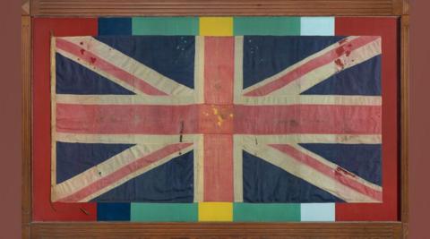 The Leiston FEPOW Memorial. It is a World War Two-era faded union jack which has been pinned to a background painted in the colours of the medal the Pacific Star: red, blue, green, yellow and white stripes and encased in a wooden frame. 