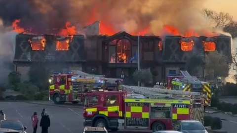 A large building is on fire with its roof collapsed. Clouds of smoke are billowing upwards. Three fire engines are parked in front of the building and a small group of people are watching on.
