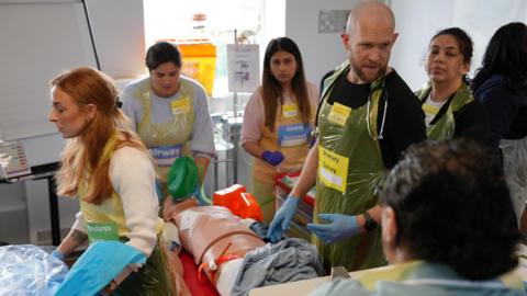 A medical training exercise. Several people wearing surgical gloves and aprons are working on a dummy patient.