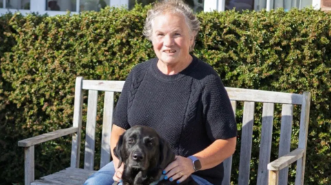 Woman in black jumper sat on bench with black dog.