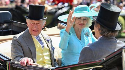A picture of Queen Camilla, wearing a light blue outfit, and King Charles, who is wearing a yellow waistcoat and grey jacket, arriving by carriage. The back of Prince Saud bin Khalid bin Abdullah bin Abdulrahman Al Saud is also pictured.
