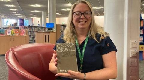 A woman with long blonde hair and glasses, wearing a blue shirt and with a green lanyard is smiling at the camera while holding a copy of the overdue hardback. She is sitting in a red leather chair in the library.