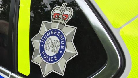 A Humberside Police logo on the side of a police car. It is a silver star with a crown at the top with ER and Humberside Police in a circle in the middle. You can see the yellow high vis on the police car.