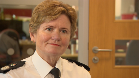 Maggie Blyth, in her uniform, at a police station. She is slightly smiling as she looks off-camera. She has short gingery blonde hair, clear blue eyes, and is wearing gold earrings. A corporate pine door with glass panels can be seen behind her in an office setting.