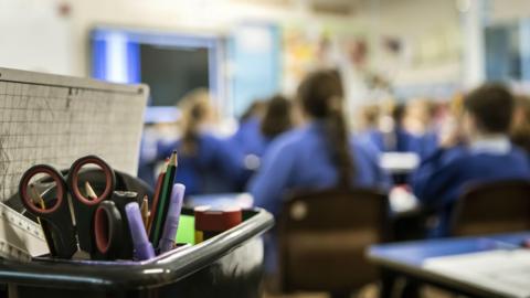A blurred picture of a classroom with a stationery kit in focus. The backs of children sitting at desks can be seen from behind, they are out of focus.