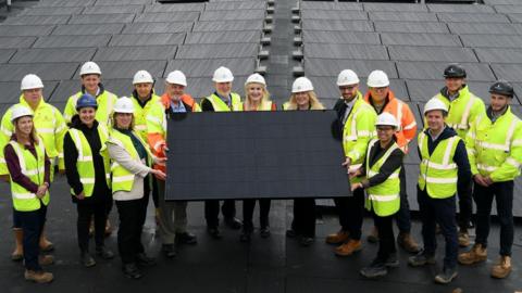 People wearing hi-vis jackets hold and stand around a solar panel on the school's roof