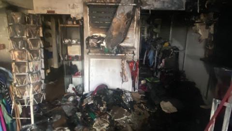 A badly burnt shop with lots of damaged goods on the floor. There is CD rack on the left, a burned material hanging from the ceiling and racks of clothes to the right.