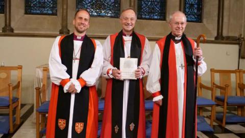 Three people stand next to each other in robes that are white, black and red. The new Bishop of Exeter Mike Harrison is stood in the middle smiling and holding a piece of paper, and the Archbishop of Canterbury is stood on his right.