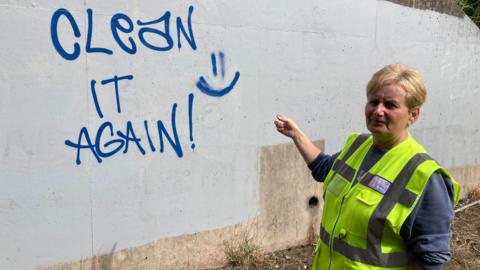 A woman in a yellow high-vis sleeveless jacket, pointing at a concrete wall which  is covered in graffiti - the graffiti spells out the message "clean it again" in blue capital letters.