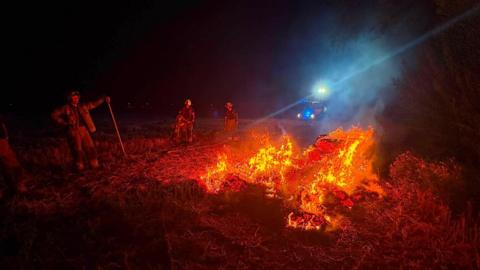 Firefighters stand in front of a group of hay bales on fire. There is white smoke rising into the black night sky.