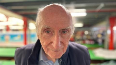 Don Cameron smiles at the camera from the ground floor of his Bedminster balloon factory with cutting tables in the background. He is an elderly man with white hair and blue eyes.