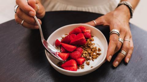 A person takes a spoonful of yoghurt, granola and strawberries from a bowl. Both of their hands are visible and they are wearing silver rings.