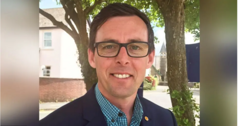 Darren Price yn edrych tuag at y camera gyda choed ac adeilad gwyn yn y cefndir. Mae ganddo lygaid glas, gwallt brown ac yn gwisgo sbectol.