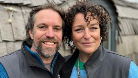 A man and a woman smiling and standing with their arms around each other in front of a cabin, the man has a beard and wears a grey bodywarmer and blue jumper. The woman has short, curly dark hair and is wearing a blue and green striped jumper and the same grey bodywarmer.