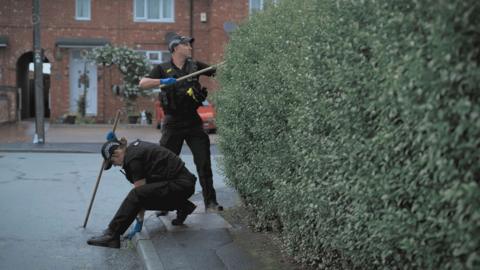 Two uniformed police officers search on a residential street. There is a large bush on the right hand side
