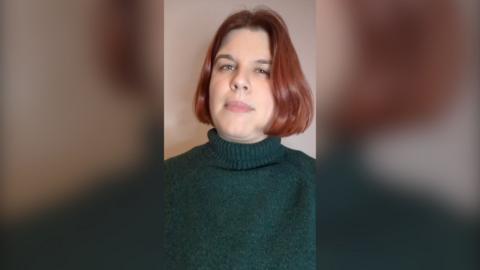 Laura Cress has short red hair and is wearing a green jumper.