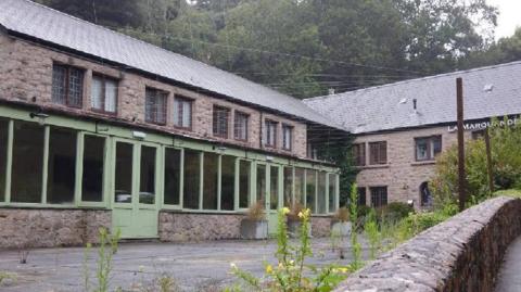 A photo which shows the disused building. The building is made of brown-grey brick and has green doors and windows. There are trees behind it with green on them.