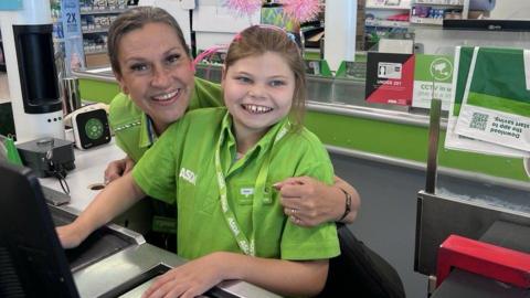 A young girl wearing an Asda supermarket uniform sits at a checkout with a woman.