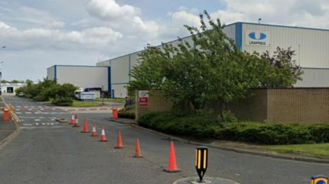 A grey and blue factory building sits on the right hand side of a road. A wall covered by trees and bushes can be seen in front of it, to the right. There are orange traffic cones laid out on the road.