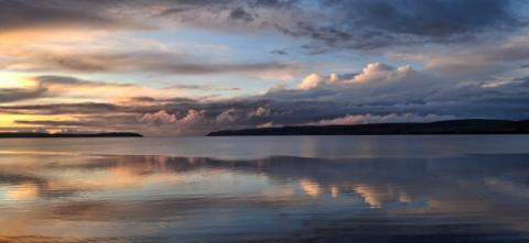 Calm coastal water reflecting a dramatic sunset sky, with soft orange and blue tones and layered clouds stretching across the horizon