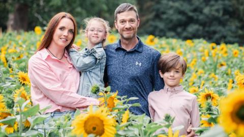 A family of four poses for a picture in the middle of a sunflower field. A woman on the left is holding a little girl up. A man is standing next to them with a boy just in front of him.