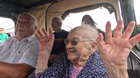 Kay White waves her hands in the air as she is driven around in a World War Two truck. She is sat next to a man wearing a white short-sleeve shirt and in front of a man wearing a black t-shirt and a woman.