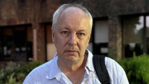 A man with short white hair wearing a blue and white shirt. He has the strap of a black bag over one of his shoulders and is standing in front of a building with bushs as its base.