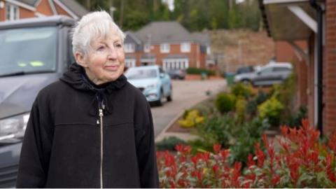 Irene Taylor looks out of the shot, has grey hair, and wearing a black hoodie. She stands in front of a flower bed and cars are behind her on the housing estate