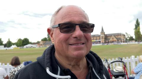 A 70-year-old man wearing sunglasses and a black hoodie smiles as he stands in front of a cricket pitch